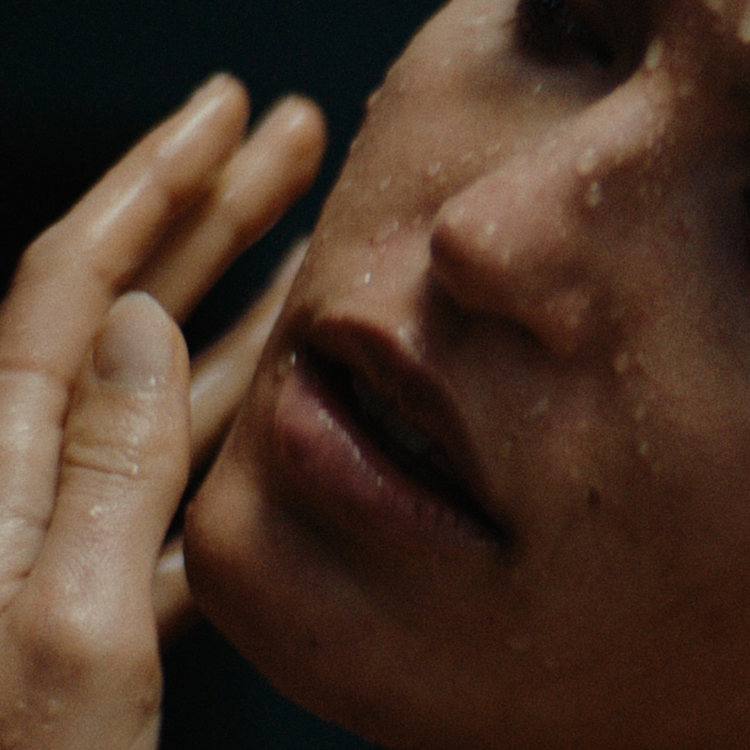 Tahi close-up of a person's face with a hand near their mouth against a dark background.